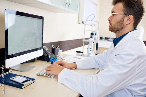 Portrait of  doctor using PC sitting at desk in office and typing on keyboard  looking at blank white screen