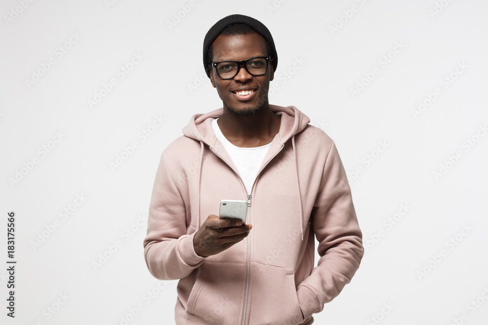 Closeup portrait of young African American man in casual clothes and spectacles pictured isolated on white background holding cellphone in hand looking at camera with open smile, browsing or texting Closeup portrait of young African American man in casual clothes and spectacles pictured isolated on white background holding cellphone in hand looking at camera with open smile, browsing or texting