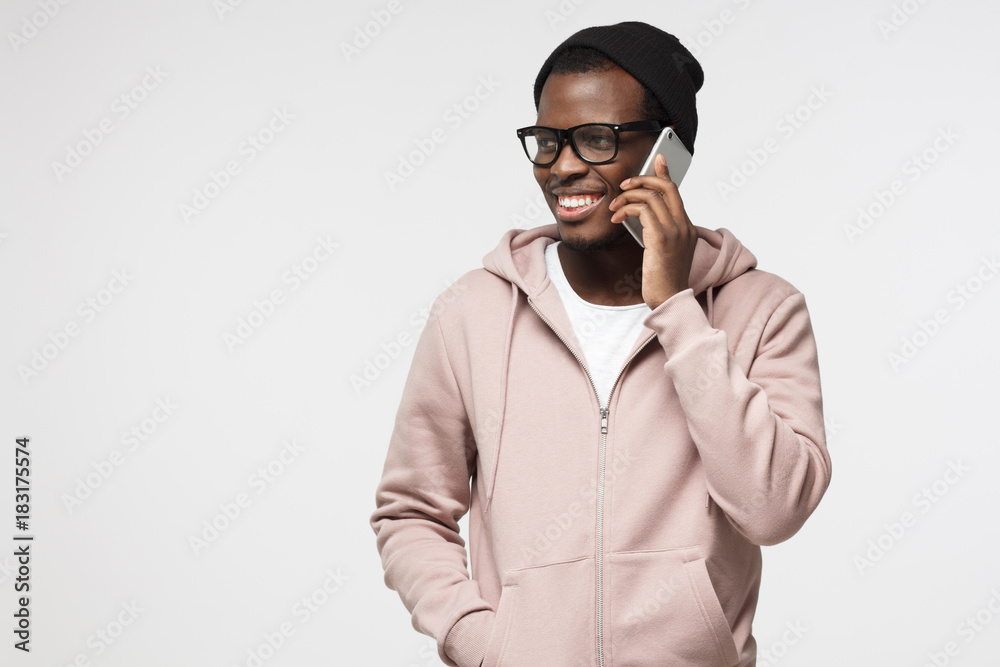 Horizontal shot of young African American man in casual clothes and glasses pictured isolated on white background looking sidewards while talking to friend on cellphone with satisfied relaxed smile