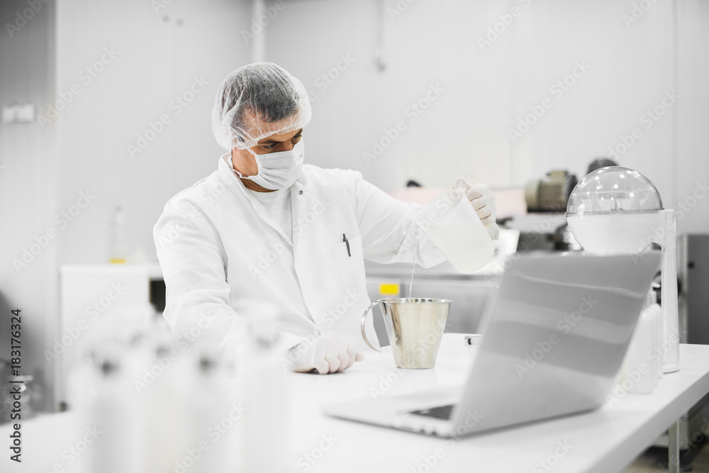 Modern professional scientist sitting at the table and pouring liquid from one flask to another at cosmetic fabric laboratory. Modern professional scientist sitting at the table and pouring liquid from one flask to another at cosmetic fabric laboratory.