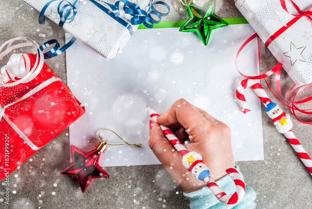 A gray table with a greeting sheet, Christmas decorations, a cup of hot chocolate and pen in form of candy cane. Girl writing, female hand in picture, copy space top view snow effect