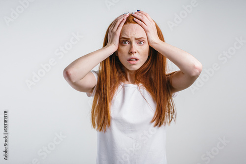 Closeup portrait of emotional redhead girl on white background with strong expression of fear, round eyes, open mouth and hands pressed to top of head, showing astonishment and shock