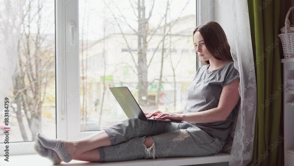 Attractive young woman sitting on a window sill and using a laptop Attractive young woman sitting on a window sill and using a laptop