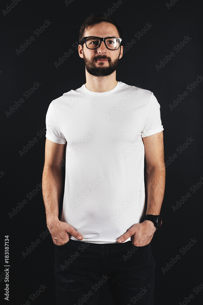 Young bearded man wearing in white t-shirt with his hands in jeans pockets on a dark background
