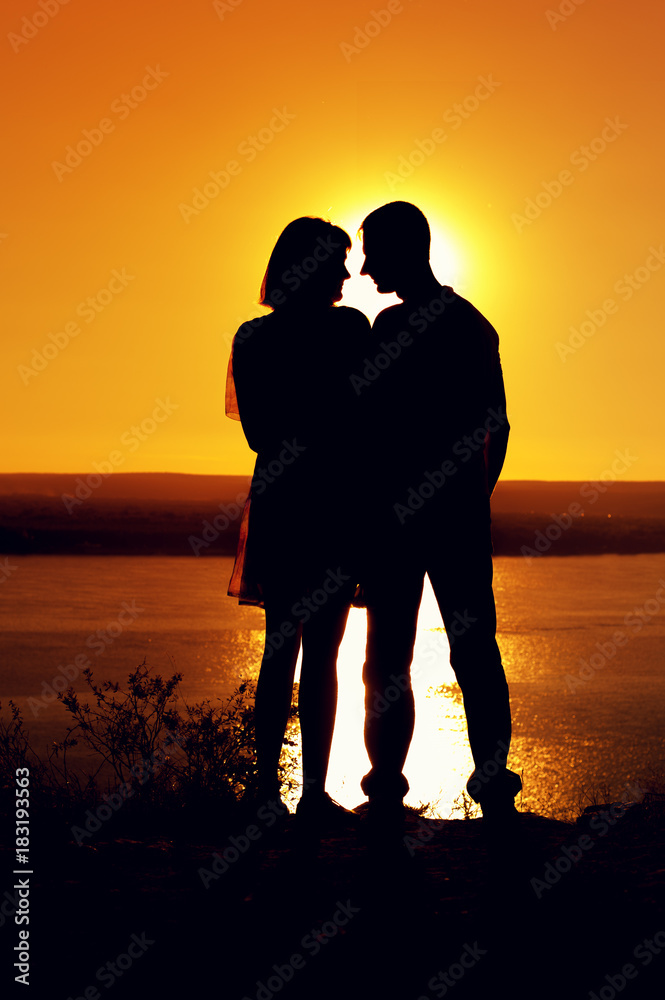 Silhouettes of young man and woman holding each other against the background of sea in sunset at the quiet sunny evening