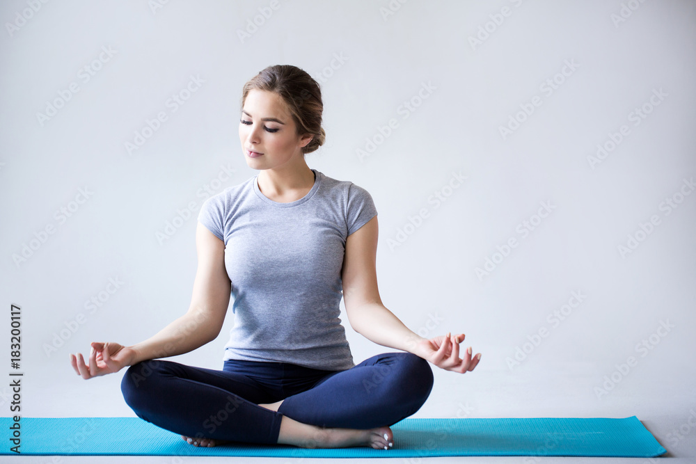 Beautiful young woman in sportswear practicing yoga while sitting in lotus position on gray background.