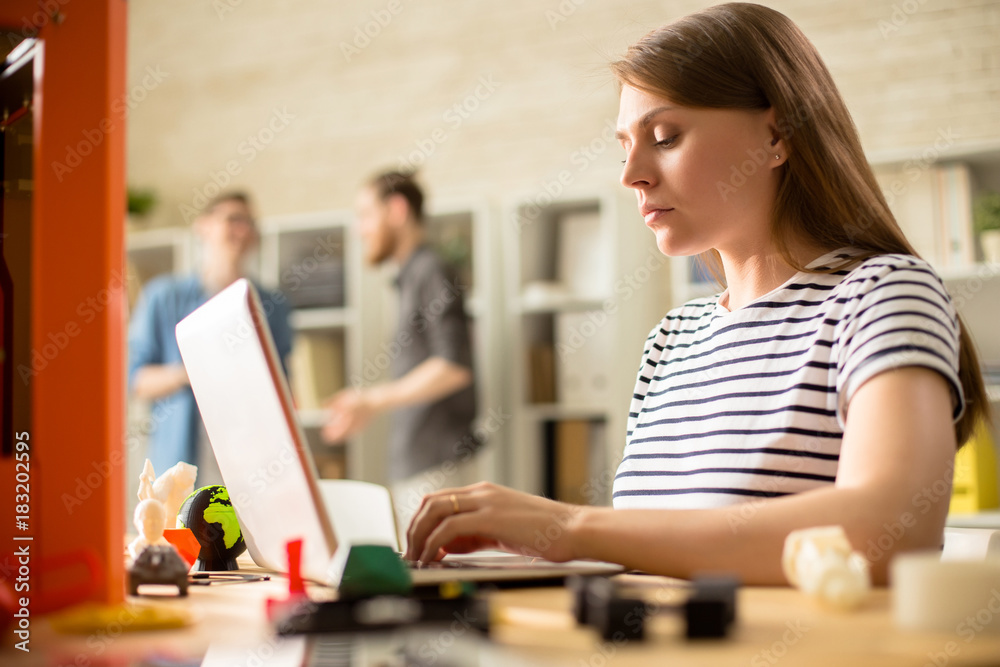 Side view portrait of pretty young woman working with 3D printer operating it via laptop in creative design studio Side view portrait of pretty young woman working with 3D printer operating it via laptop in creative design studio