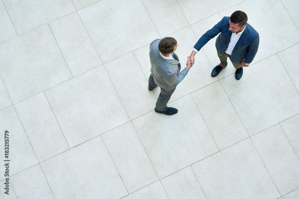 Top view of two business people shaking hands standing on tiled floor in hall of modern office building, copy space - obrazy, fototapety, plakaty 