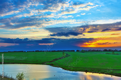 Colorful landscape on the river bank in spring, orange sun hiding behind blue clouds in a beautiful sky. Sunset on the lake in Russia.
