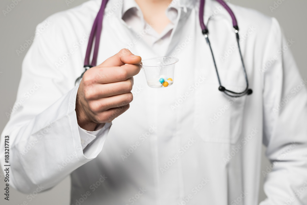 close up of Smiling doctor offering a cup of pills or drugs on gray background. Daily medicine, dose or vitamins concept close up of Smiling doctor offering a cup of pills or drugs on gray background. Daily medicine, dose or vitamins concept