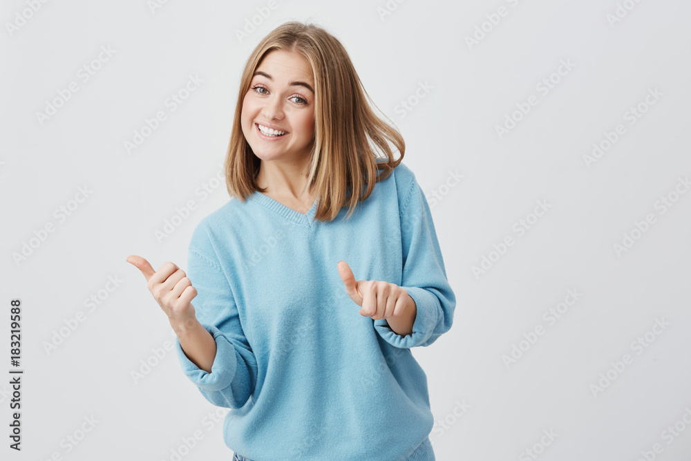 Emotional happy young caucasian female with fair hair dressed in blue clothes giving her thumbs up, showing how good a product is. Pretty girl smiling brodly with teeth. Gestures and body language Emotional happy young caucasian female with fair hair dressed in blue clothes giving her thumbs up, showing how good a product is. Pretty girl smiling brodly with teeth. Gestures and body language