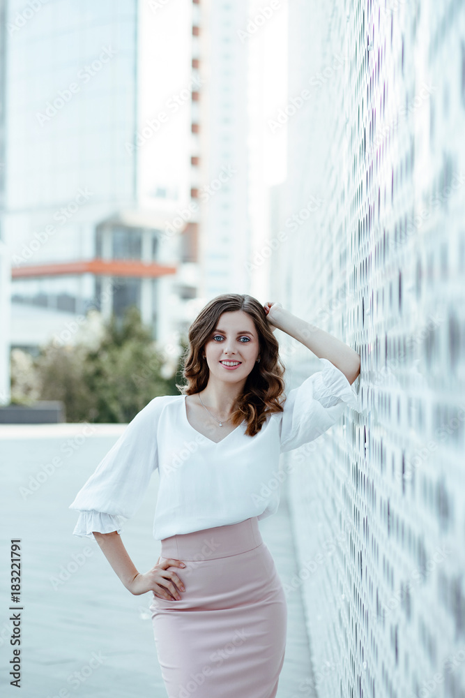 Office style. A business portrait of the young woman in a white blouse and a pink skirt against the background of city architecture.