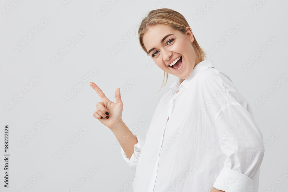 Advertising concept. Good-looking young woman with blonde hair wearing casual clothes, joyfully smiling withteeth, standing against gray studio wall with copy space for promotional content Advertising concept. Good-looking young woman with blonde hair wearing casual clothes, joyfully smiling withteeth, standing against gray studio wall with copy space for promotional content