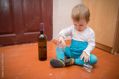 a little baby in dirty, torn clothes plays with a broken glass on the wooden floor, next to it there is a bottle with alcohol