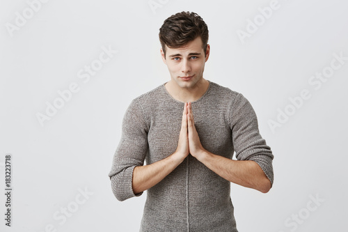 Handsome young man with dark hair pressings palms together and looking at camera with his blue eyes, begging for forgiveness or asking to make his dreams come true. Human emotions and feelings photo