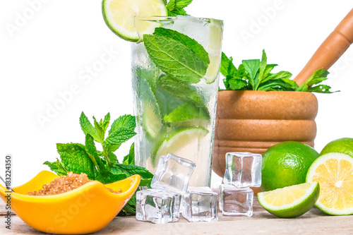 Mojito cocktail with ingredients. Fresh lime, mint leaves and ice cubes, morter with mint and brown sugar on a wooden background. Refreshing alcohol drink. Isolated on white background..