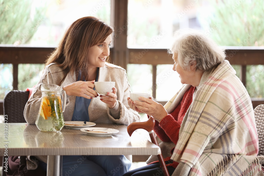 Senior woman and young caregiver drinking tea at table in cafe Senior woman and young caregiver drinking tea at table in cafe