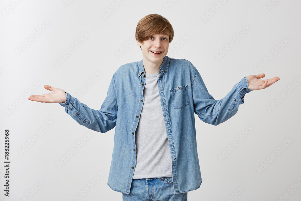 Shot of smiling young male wears denim clothes with fair hair gestures with hands, says: who cares, I don`t know anything, has indignant expression, poses against gray studio background. Shot of smiling young male wears denim clothes with fair hair gestures with hands, says: who cares, I don`t know anything, has indignant expression, poses against gray studio background.