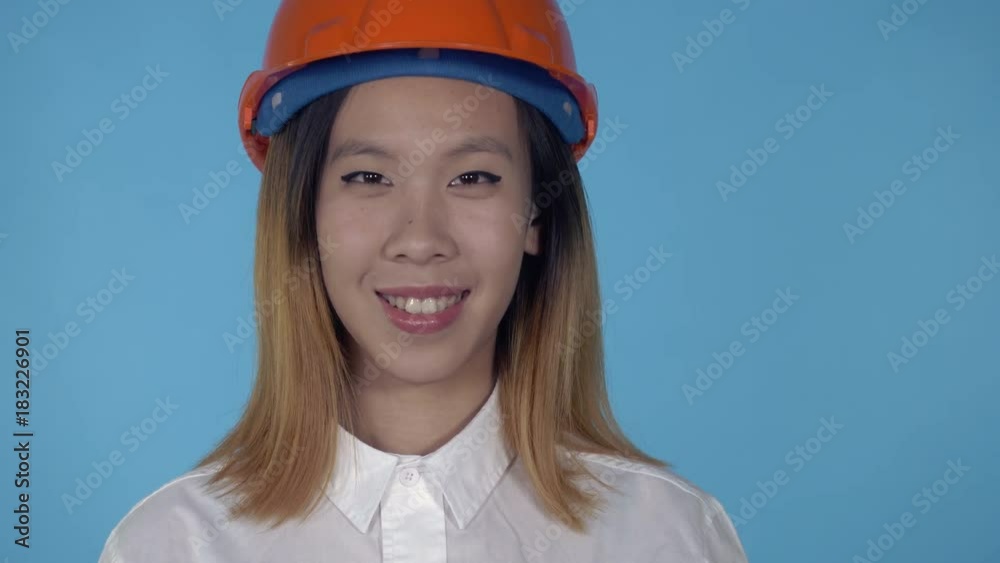 close up portrait young asian architect wearing orange hard hat shows symbol ok on blue background in studio. attractive millennial girl looking at the camera
