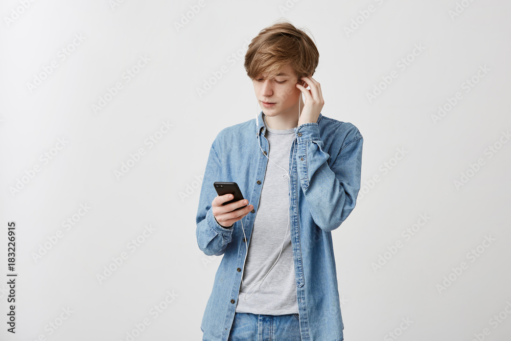 Indoor shot of young caucasian man with fair hair dressed in denim shirt resting after classes at university. Guy listening music in white earphones, using music app on smartphone. Indoor shot of young caucasian man with fair hair dressed in denim shirt resting after classes at university. Guy listening music in white earphones, using music app on smartphone.