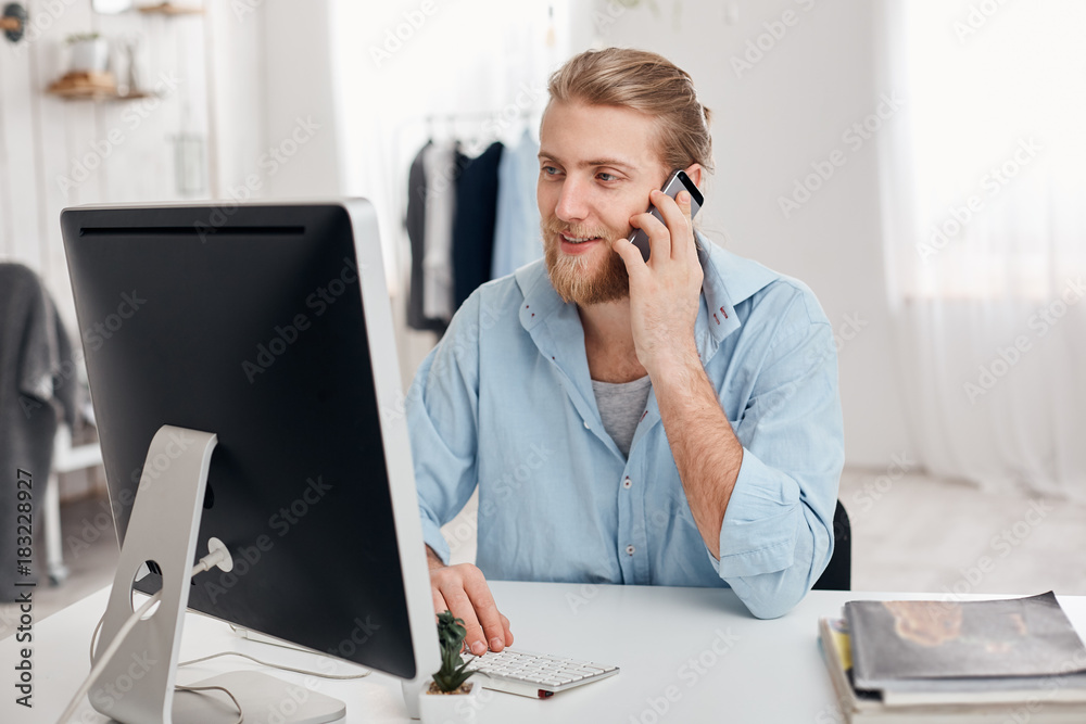 Bearded skilled young fair-haired copywriter works on new article, types on keyboard, has phone conversation, discusses new project with businesspartner. Successful businessman has important call. Bearded skilled young fair-haired copywriter works on new article, types on keyboard, has phone conversation, discusses new project with businesspartner. Successful businessman has important call.