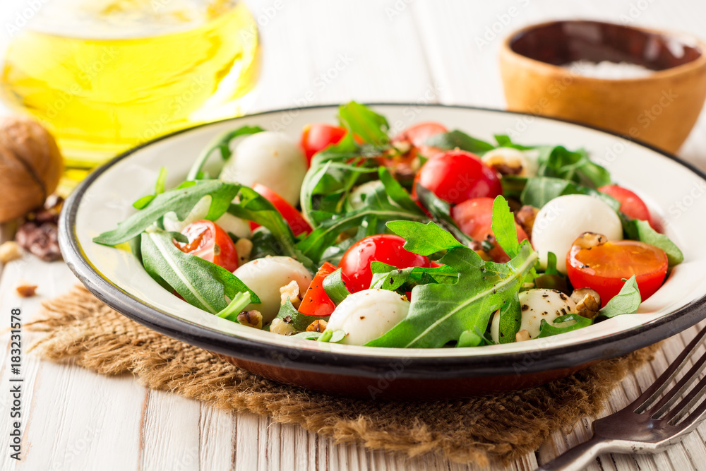 Fresh salad with arugula, cherry tomatoes, mozzarella cheese and walnuts on white wooden background.