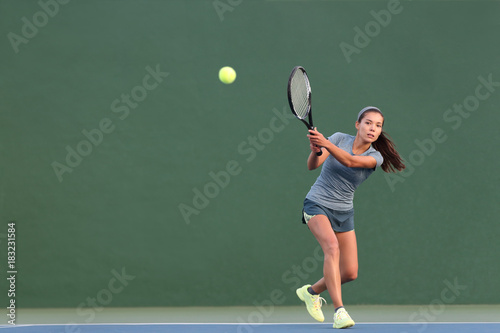 Tennis playing woman hitting ball on green hard court. Asian athlete girl returning serve with racket wearing skort and shoes.