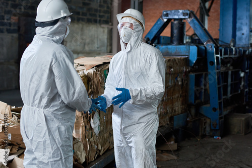 Portrait of two workers wearing biohazard suits communicating in industrial warehouse of modern waste processing plant against recyclable cardboard in background, copy space
