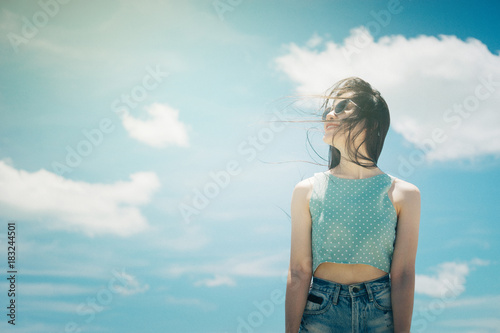 Portrait of a cute asian teenager young woman stand against blue sky and cloud background. wearing sunglasses. wind blows her hair. filtered image to vintage tone. light effect added. hipster concept