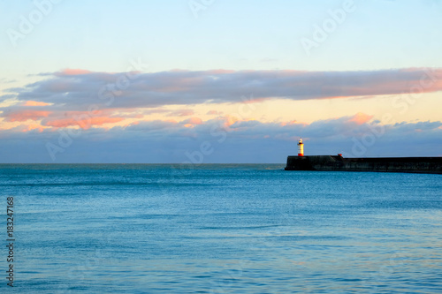 light house at end of sweeping harbour wall on left hand side calm blue sea in foregroundsunset sky