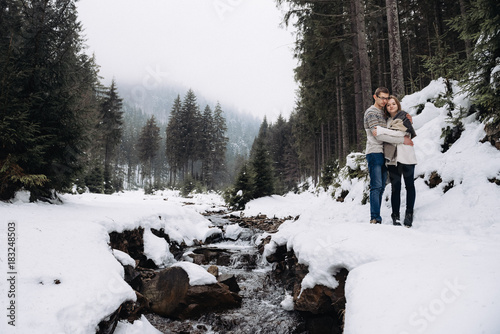 Young beautiful couple staying near the winter mountain river, hug and smile photo