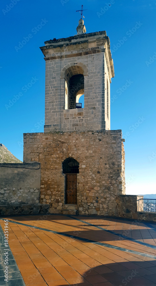Old stone belltower with wooden door and metal cross. Blue clear sky.