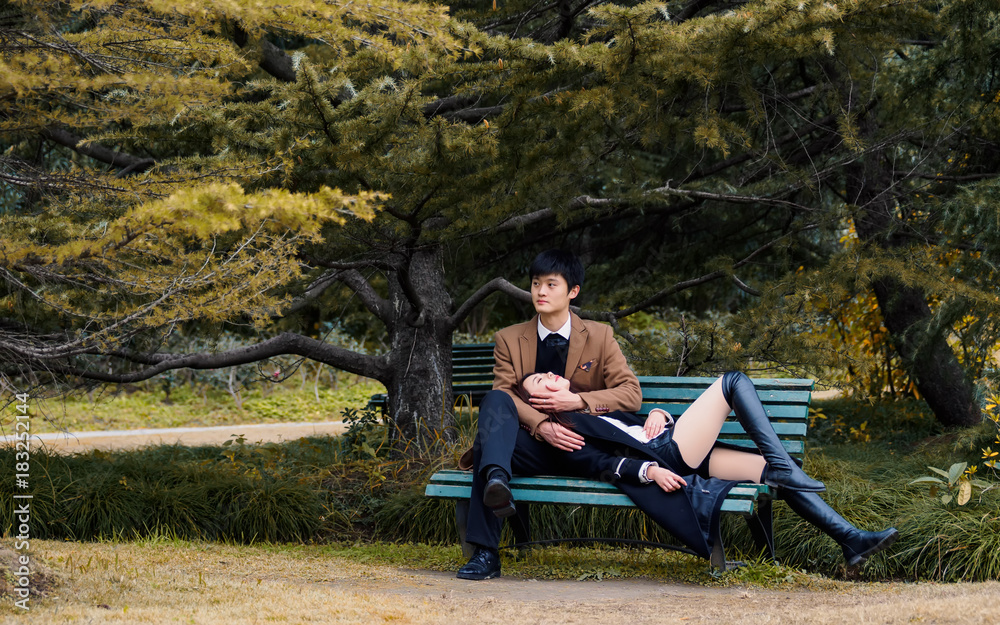 Portrait of couple of Chinese young people sit in bench in autumn park, girl lying on the thigh of her boyfriend, lover concept.