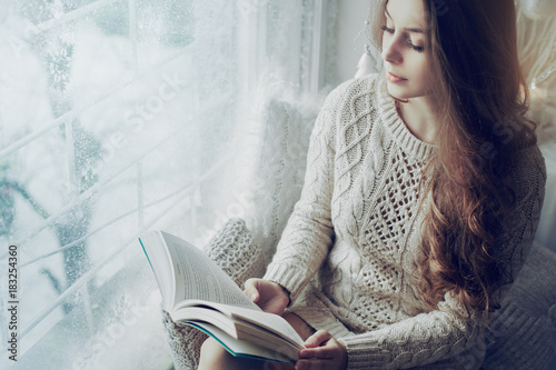 Young beautiful girl wearing knitted dress sitting on windowsill, holding and reading book. Day light. Christmas, New year, winter holidays concept. Copy space for text. 