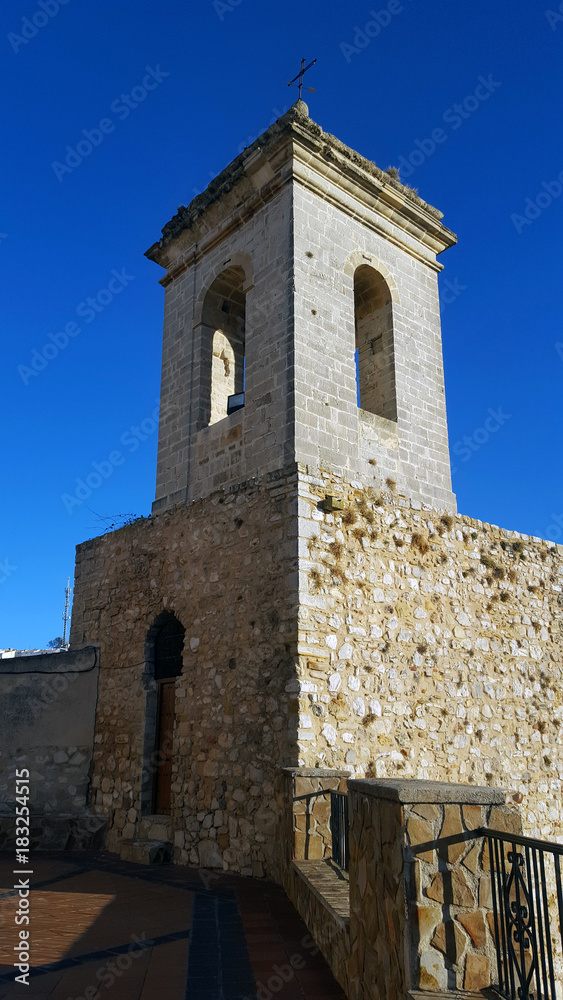 Old stone belltower with wooden door and metal cross. Blue clear sky.