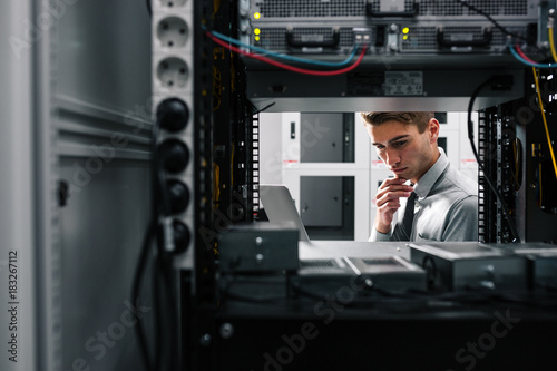 Portrait of modern young man holding laptop standing in server room working with supercomputer