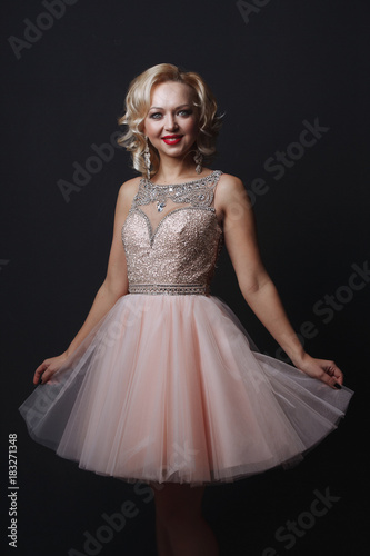 Portrait of a beautiful sensual young blonde woman with blue eyes in pink dress with rhinestones, laughing, on a dark background in the Studio