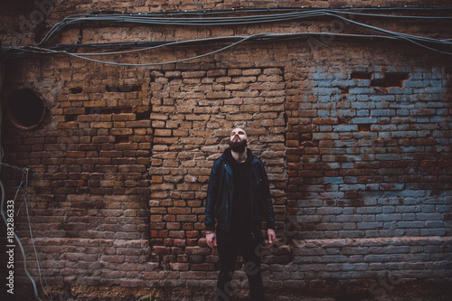 Modern young bearded man in black style clothes posing against brick wall.