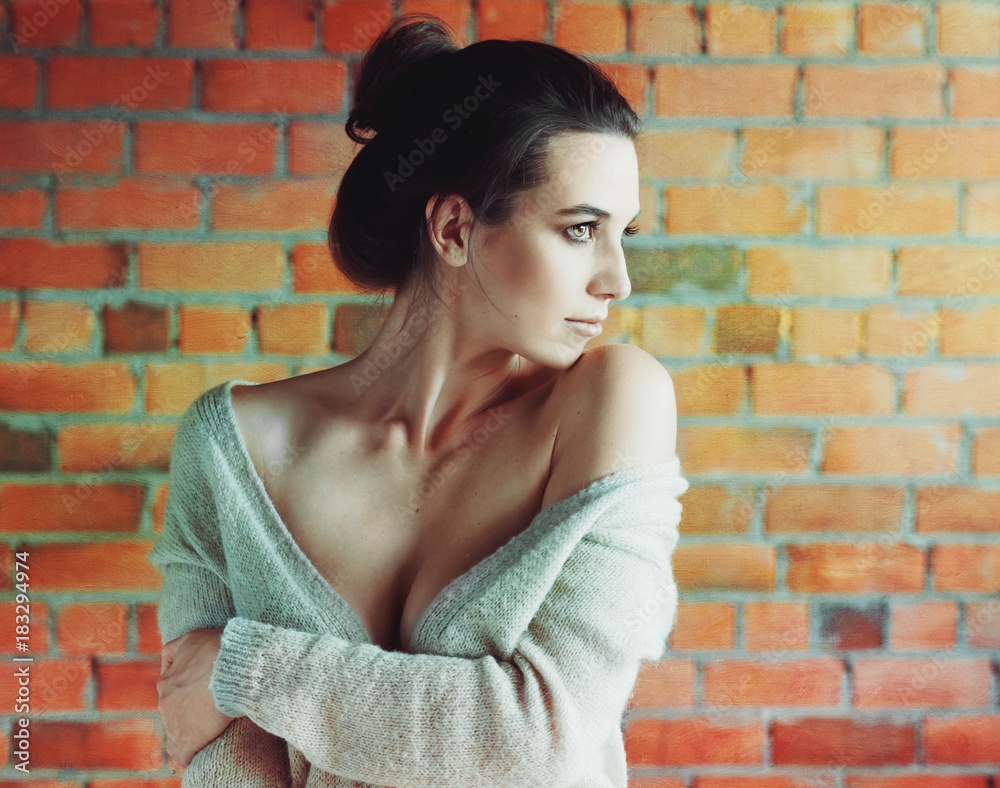 Stylish brunette woman in white sweater posing near red brick wall