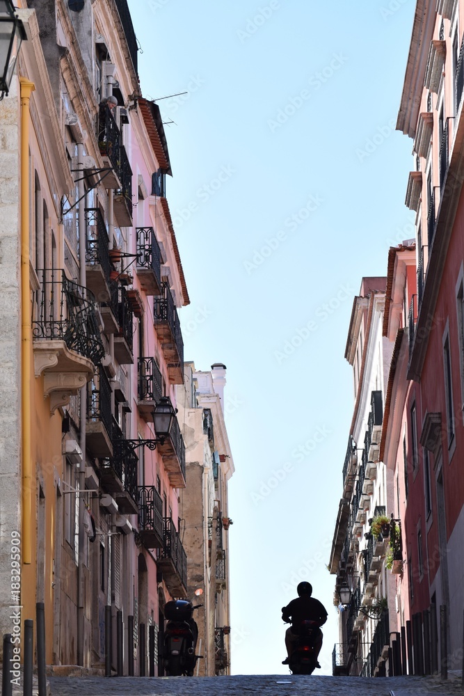 Fototapeta premium Straßenschlucht in Lissabon, mit blauem Himmel und Rollerfahrer
