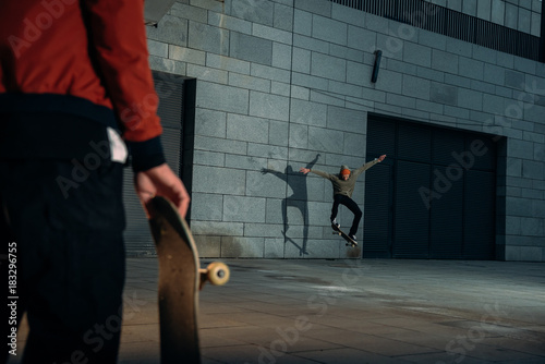 cropped shot of man standing with skateboard while his partner doing trick on background