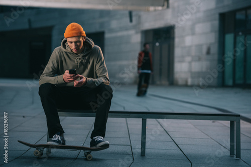 Skateboarder in modern streetwear sitting on bench and using smartphone photo