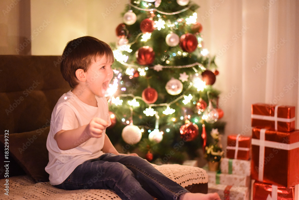 Happy boy hold burning sparkler and smiling In front of Christmas tree. Young boy holding burning sparkler. Winter, holiday, vacation, Christmas Happy boy hold burning sparkler and smiling In front of Christmas tree. Young boy holding burning sparkler. Winter, holiday, vacation, Christmas