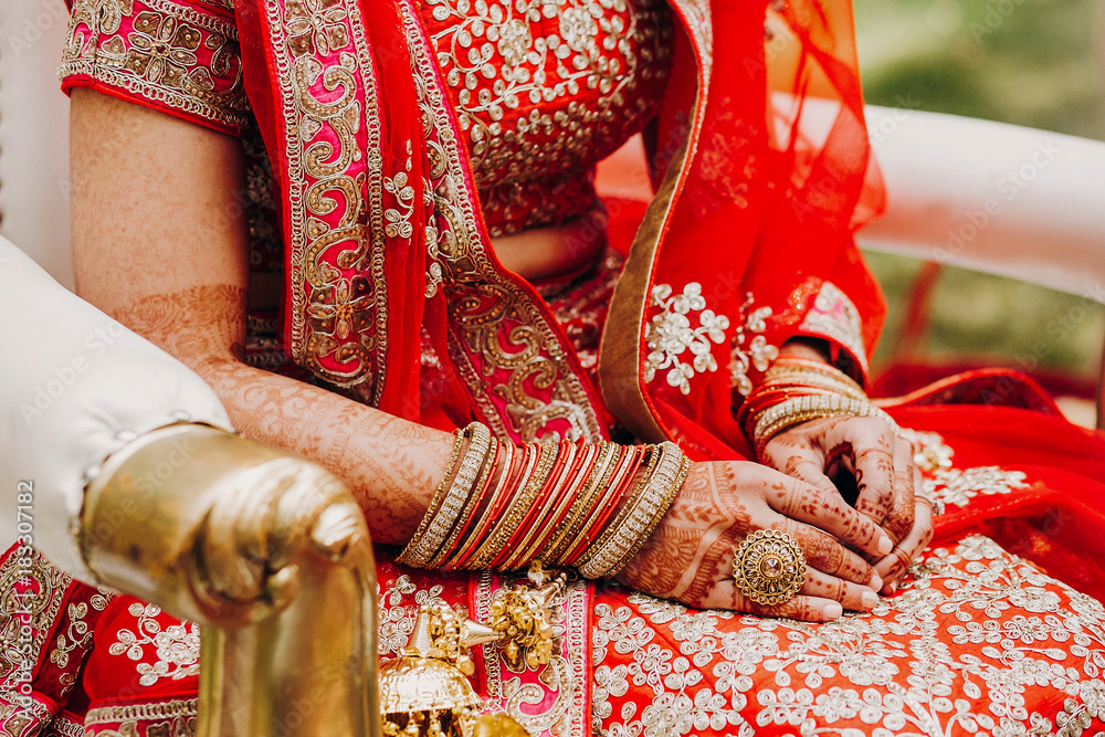 Indian bride in red lehenga embroidered with gold holds her hands with henna tattoos on the knees during wedding ceremony
