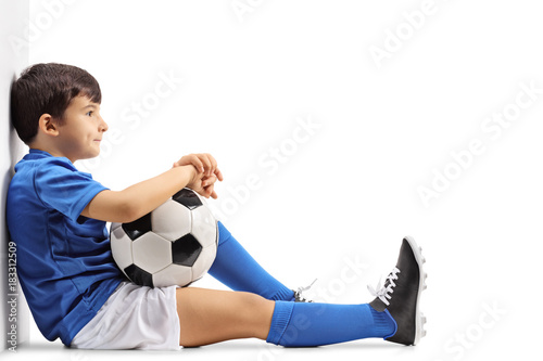 Pensive little footballer sitting on the floor and leaning against a wall