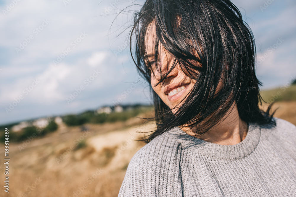 Horizontal cropped portrait of beautiful smiling woman wearing sweater being playful with windy hair and carefree posing on sunlight sky and nature background. Travel, people and lifestyle concept.