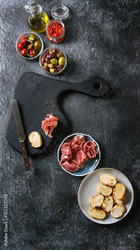 Ingredients for making tapas or bruschetta. Crusty bread, ham prosciutto, sun dried tomatoes, olive oil, olives, pepper with wooden serving board over dark texture background. Top view with space
