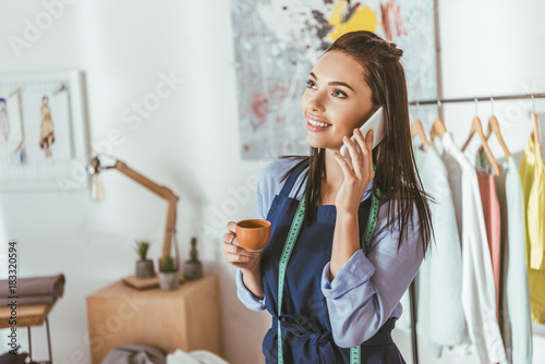 smiling seamstress talking by smartphone and holding cup of tea
