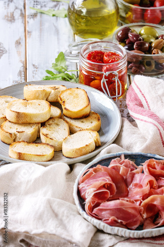 Ingredients for making tapas or bruschetta. Crusty bread, ham prosciutto, sun dried tomatoes, olive oil, olives, pepper, greens on plates over white plank wooden table. Close up