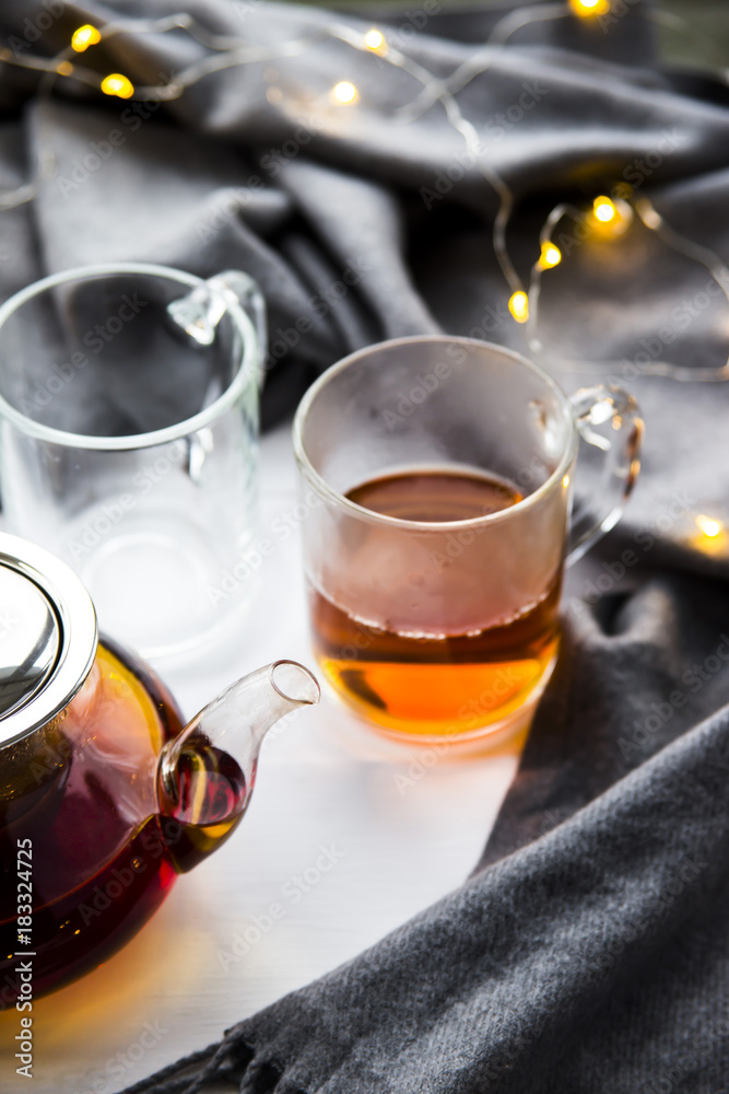 Black transparent teapot and cups with black tea on a wooden table next to a gray scarf and lights. Winter home concept Black transparent teapot and cups with black tea on a wooden table next to a gray scarf and lights. Winter home concept